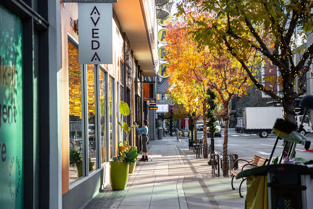 A city sidewalk lined with shops, autumn trees with yellow leaves, parked bikes, and a person standing outside a storefront window. - Atelier Studio, San Jose, CA