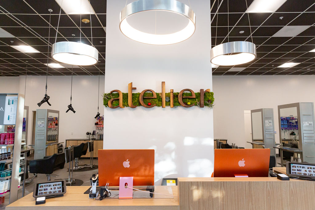 Reception area of a salon with two orange iMac computers, a sign reading "atelier" on the wall, and modern lighting overhead. - Atelier Studio, San Jose, CA