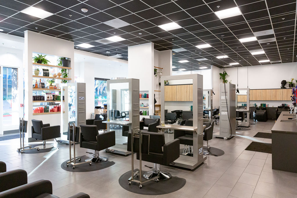 Modern hair salon interior with multiple styling stations, black chairs, mirrors, shelves with hair products, and large windows providing natural light. - Atelier Studio, San Jose, CA