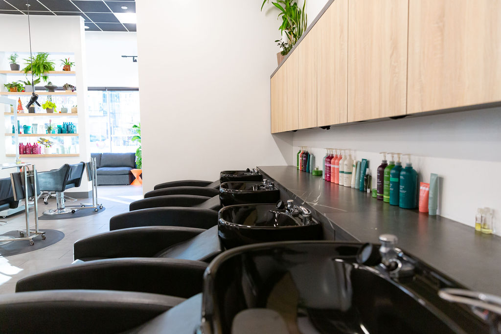 A row of black salon washbasins with hair products on the counter, wooden cabinets above, and seating area visible in the background. - Atelier Studio, San Jose, CA