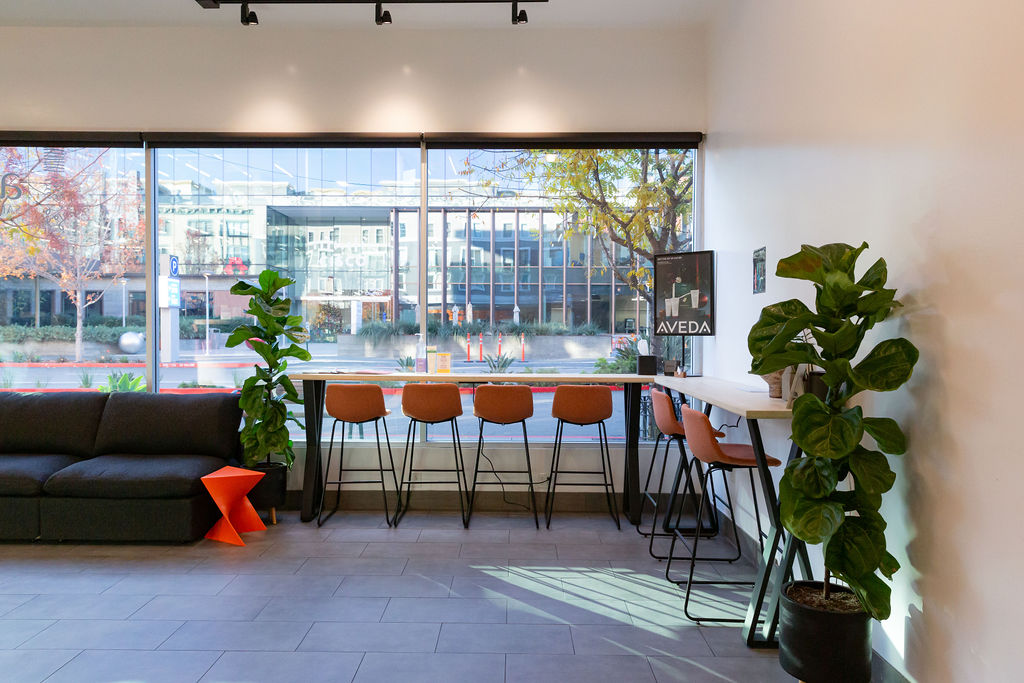 Modern lounge area with high chairs at a window counter, potted plants, a black sofa, and a cityscape visible through large windows. - Atelier Studio, San Jose, CA