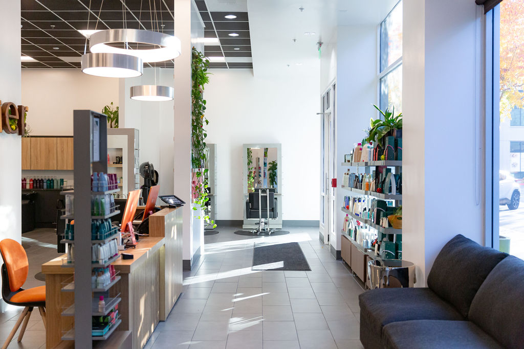 Modern salon interior with product shelves, reception desk, styling stations, gray sofa, and large windows letting in natural light. - Atelier Studio, San Jose, CA