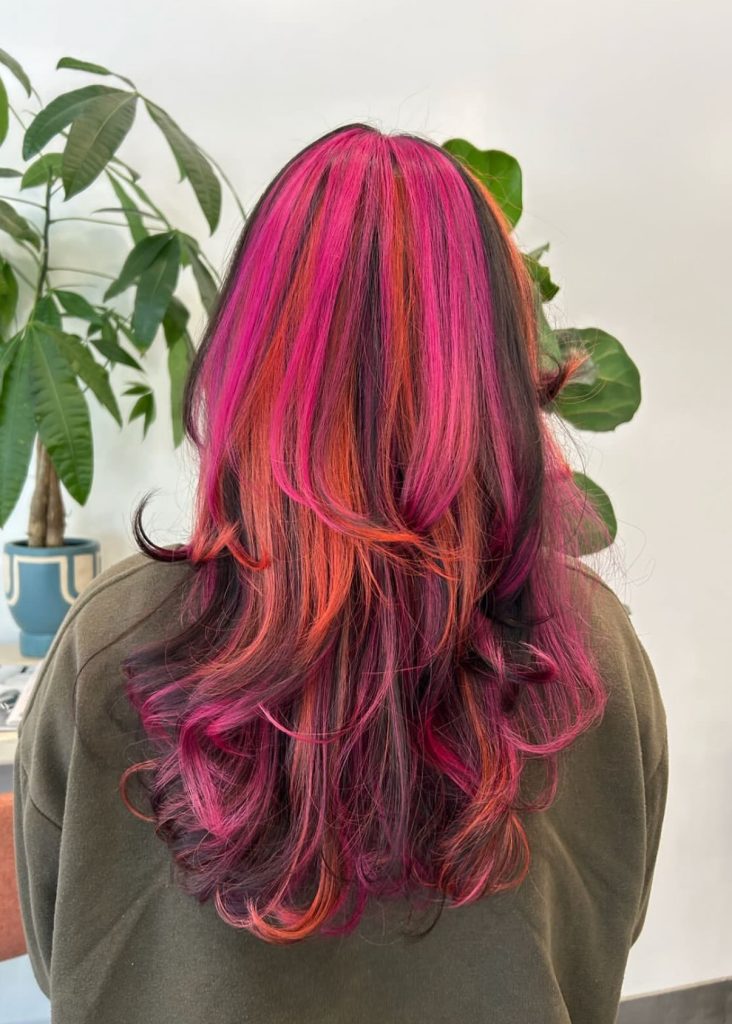 A person with long, wavy hair dyed in bright pink, orange, and purple tones, photographed from behind, with a potted plant in the background. - Atelier Studio, San Jose, CA
