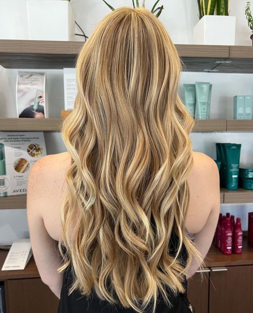 A woman with long, wavy, blonde hair stands facing away in a salon, with shelves of hair products and plants in the background. - Atelier Studio, San Jose, CA