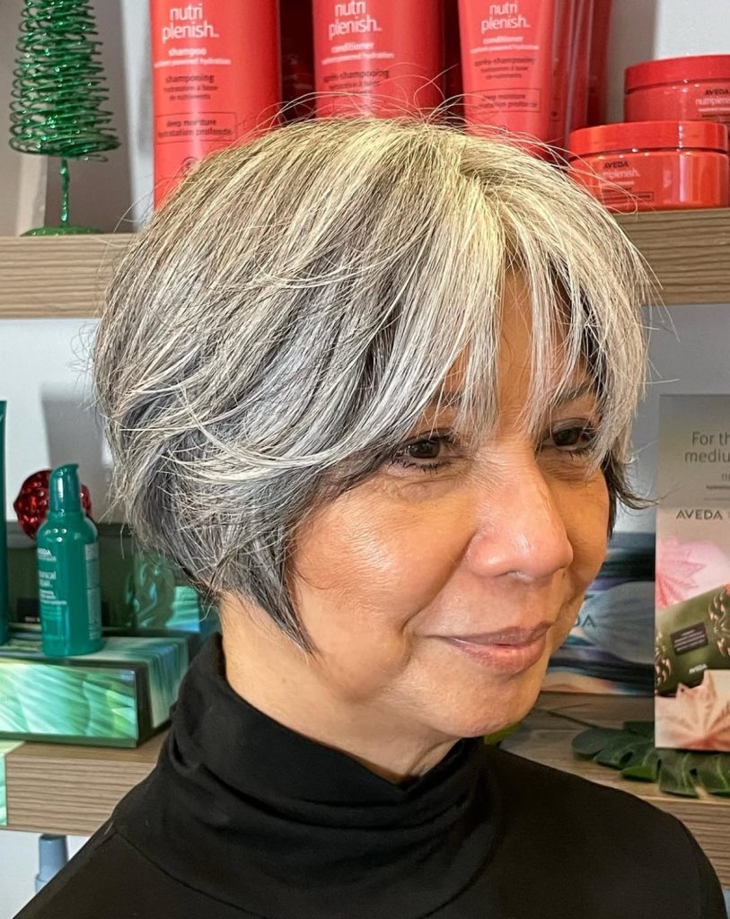 Woman with short, layered gray hair and side-swept bangs, wearing a black turtleneck, standing in a salon with hair products on shelves behind her. - Atelier Studio, San Jose, CA
