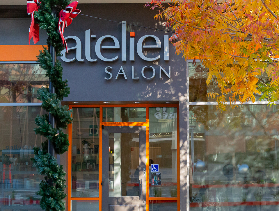 Shopfront of Atelier Salon with orange and grey exterior, a glass door, accessibility sign, holiday wreath, and autumn tree branches. - Atelier Studio, San Jose, CA